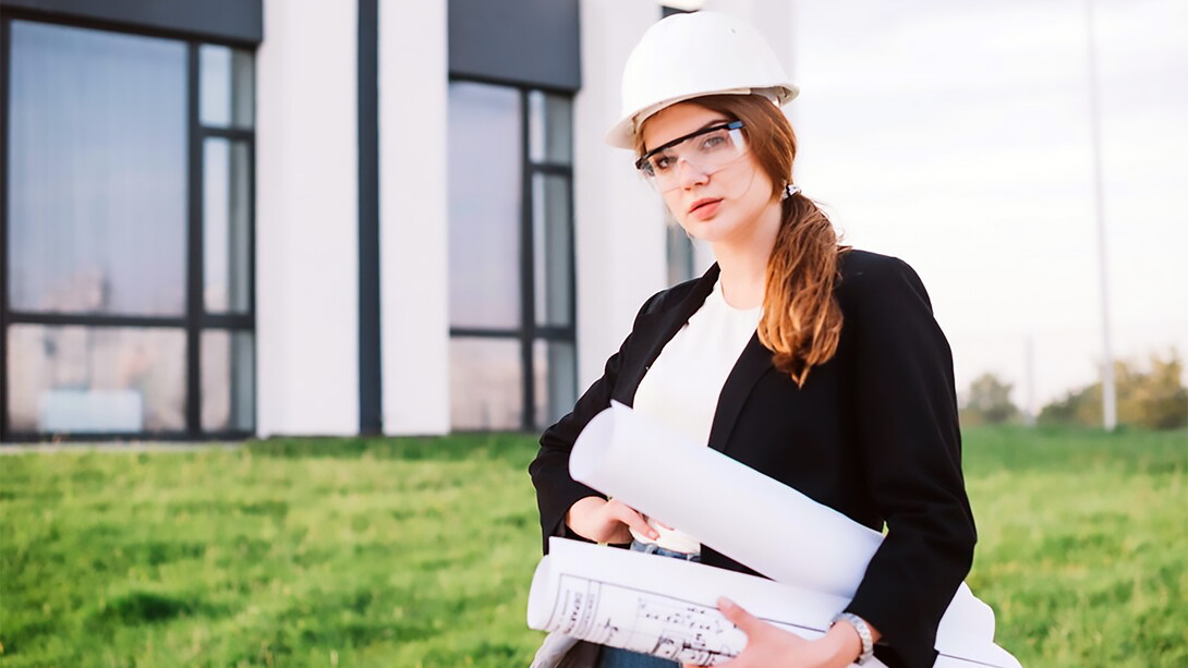 A female architect stands confidently in front of a building, holding architectural plans in her hands