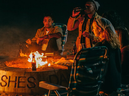 Friends surrounding a firepit on the beach, device-free, having fun and drinking beers