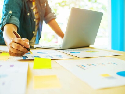 Primo piano di una persona che scrive o annota dei grafici su documenti con una penna, mentre lavora al laptop
