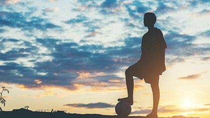 A young footballer silhouetted against the evening sky with a ball at their feet, representing dreams that reach far beyond the horizon