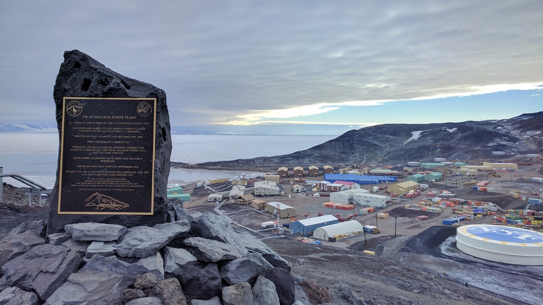 De 1962 a 1972, el reactor nuclear PM-3A operó en la Estación McMurdo. Es la única tecnología de energía nuclear de este tipo que opera en la Antártida. Este marcador conmemora su existencia. Al fondo se puede ver la Estación McMurdo. Es la comunidad más grande de la Antártida y sirve como base de investigación científica para Estados Unidos