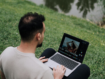 A man sits on the grass by a scenic pond in a park, using his laptop while working and enjoying the calm outdoor setting