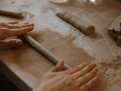 As mãos modelam uma porção de massa sobre uma mesa de madeira, dando início à construção de uma forma. Na técnica do acordelado, o movimento de enrolar a argila também é o primeiro passo para construir uma peça camada por camada