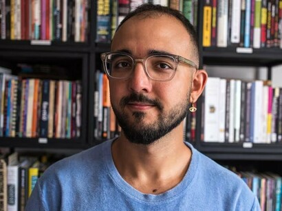 Em seu espaço de trabalho, Raphael Montes aparece entre livros e objetos ligados à cultura literária. A imagem remete à trajetória editorial do autor e à produção de obras que posteriormente chegaram ao audiovisual