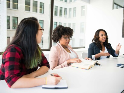 Três mulheres compartilham uma mesa em um ambiente de trabalho, em uma situação de convivência e troca. A linguagem se constrói também na relação entre sujeitos que ocupam diferentes posições na comunicação