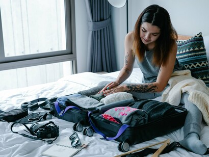 Sentada sobre a cama, uma mulher organiza roupas e outros itens dentro da mala antes de sair de casa. Preparar a bagagem com antecedência é apresentado no artigo como uma maneira de diminuir a sensação de descontrole que pode acompanhar a expectativa da viagem