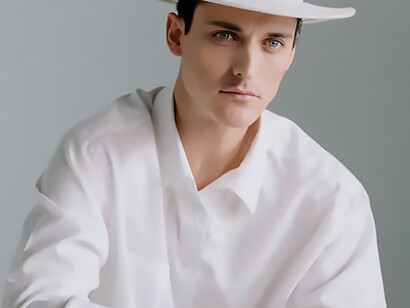 A man, dressed in a crisp white shirt and sporting a matching white hat, sits in contemplation in front of a chessboard, ready for a strategic match