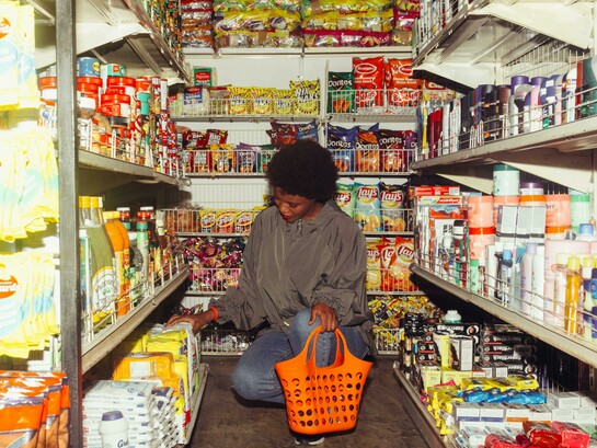 An African American woman selects products while shopping in a convenience store, carrying an orange shopping basket
