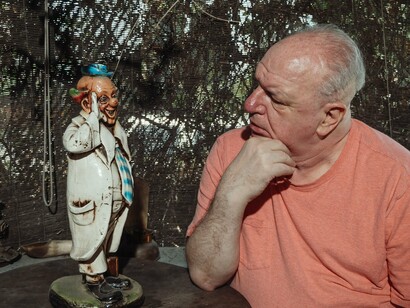 Um homem idoso observa de perto uma pequena figura de palhaço, estabelecendo uma relação silenciosa entre o observador e o personagem. A cena traduz visualmente uma das ideias centrais do artigo: o palhaço pode funcionar como um espelho simbólico das nossas próprias fragilidades, expondo aquilo que muitas vezes preferimos não reconhecer em nós mesmos