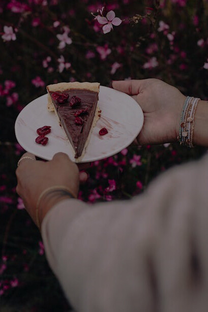 A woman elegantly poses amidst pink flowers, holding a plate adorned with a tempting chocolate cake, capturing a delightful moment of indulgence