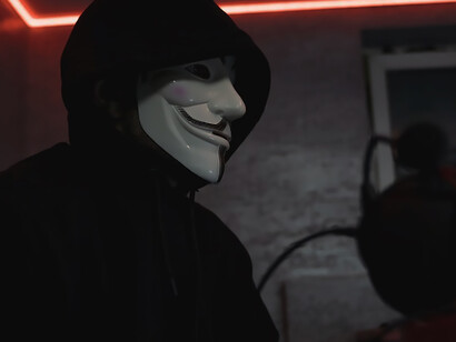 Cloaked in a Vendetta mask, a hacker illicitly operates on a computer, concealed in the shadows