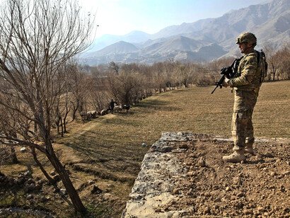A soldier stands in a field on a wall next to a line of trees, with a weapon in hand, in Afghanistan