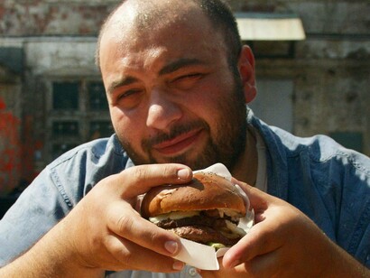 Primo piano di un uomo che tiene in mano un hamburger avvolto nella carta e guarda verso la telecamera con un leggero sorriso: "Diversi studi confermano che l’informazione diffusa sui social media segue logiche simili a quelle della pubblicità: è progettata per essere consumata rapidamente e suscitare una risposta emotiva immediata"