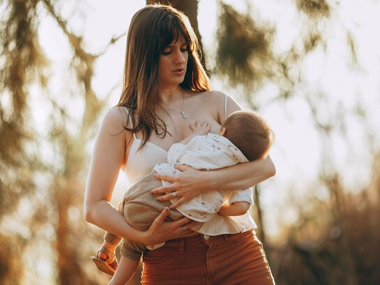 Mulher segura o bebê junto ao corpo em meio à floresta, sob a luz suave do fim de tarde. O vínculo entre mãe e filho está no centro da discussão sobre os benefícios e as características do leite materno