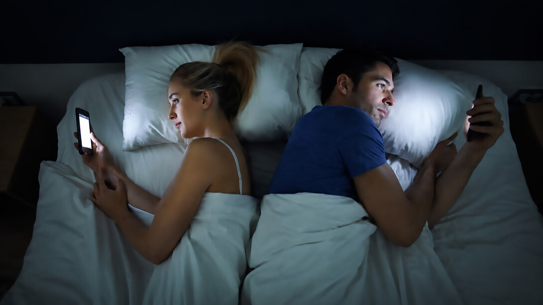 A husband and wife lie back-to-back in bed, each absorbed in scrolling through their phone