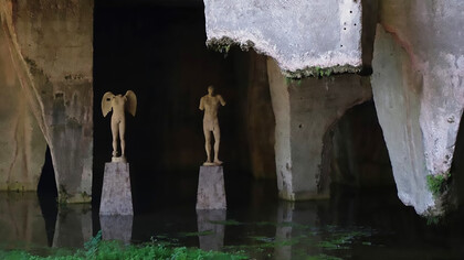 Abandoned and neglected sculptures stand in a cave beside a still body of water, remnants of a forgotten artistic legacy