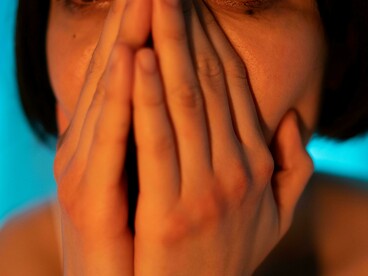 A short-haired woman in a white tank top rests her hand on her face, looking upset