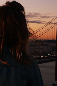 A woman stands gracefully in front of a Lisbon bridge at the enchanting backdrop of a Portuguese sunset