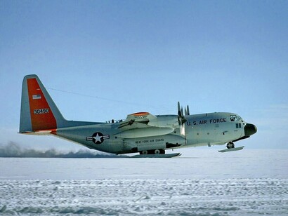 Um LC-130H Hercules decola da superfície gelada da Groenlândia, deixando um rastro de neve atrás de si. A aeronave simboliza a capacidade de alcançar e manter operações em áreas remotas do Ártico