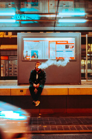A man sitting on ledge smoking during night time, as neon lights flash by