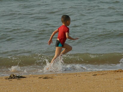 Menino correndo na beira do mar. Corri pela areia, esbarrando nas pessoas, desviando de famílias sentadas com seus guarda-sóis e crianças brincando com baldinhos. Tudo ao meu redor se tornou um borrão. O zepelim seguia em direção ao norte, e eu o seguia como se ele fosse um ímã e eu, uma lasca de metal