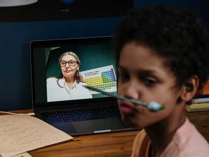 Um estudante aparece em primeiro plano diante do computador portátil, enquanto uma professora apresenta o conteúdo de uma aula online na tela. Sobre a mesa, uma atividade escolar aguarda sua atenção. A cena remete aos desafios de manter o envolvimento na aprendizagem a distância, em que estar diante da tela nem sempre significa estar plenamente atento ao conteúdo