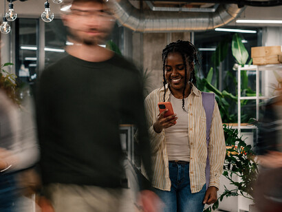 A young woman walks through a busy office corridor, absorbed in her smartphone and disconnected from the people and activity around her