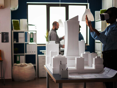Architect using VR goggles to explore a 3D plan of a foam model in a contemporary office, with an engineer wearing a headset beside an urban project model