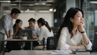 A woman sitting apart from her coworkers in a busy office, representing the emotional distance that can exist within a workplace