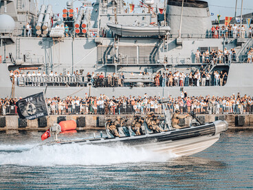 Embarcación militar de intervención rápida cruzando frente a un buque de guerra repleto de espectadores en el muelle