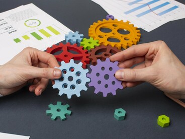 The hands arranging the gears emphasize the crucial role of human capital in driving economic activity, as labor is one of the most vital resources for production