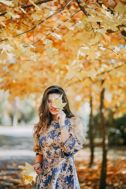 In this evocative image, a woman playfully conceals her face with a large, vibrant leaf, adding a touch of whimsy to the autumn scenery