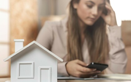 A woman holds her phone while standing behind a miniature wooden house, deep in thought