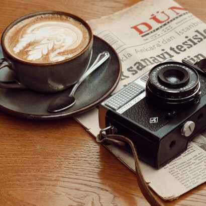 Un periódico y una cámara analógica acompañan una taza de café, evocando una época en la que el acceso a la actualidad estaba ligado a la lectura pausada y al trabajo paciente de quienes la registraban