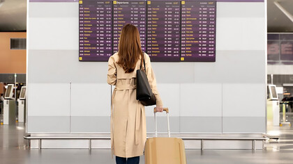 A woman traveling with baggage gazes at the departure board, representing skilled migration and brain drain as she embarks on a journey abroad for new opportunities