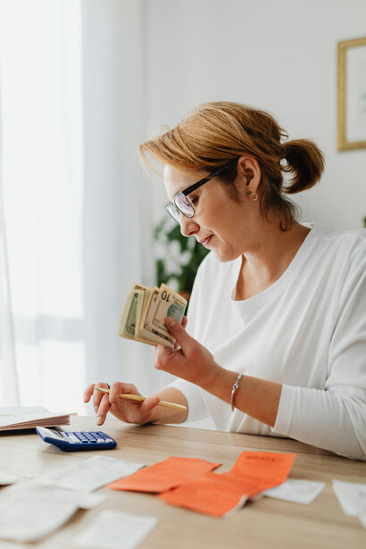 A woman using a calculator to count money and receipts on a kitchen table, managing household expense