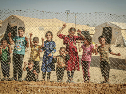 Syrian children peer through a chain lock fence at a refugee camp, their innocent faces a stark reminder of the human cost of conflict