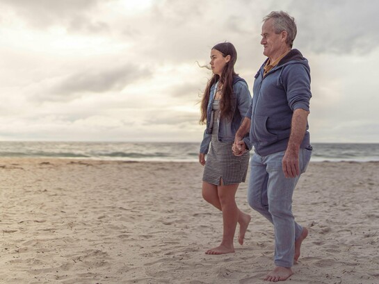 Pai e filha caminham lado a lado à beira-mar. Relações de afeto e cuidado também moldam a forma como homens envelhecem. Entre passos lentos e companhia constante, o cuidado aparece como vínculo e não apenas obrigação. Em muitos casos, são justamente as mulheres que sustentam redes emocionais e de cuidado ao longo da vida. O texto discute como masculinidade, afeto e cuidado influenciam até a expectativa de vida