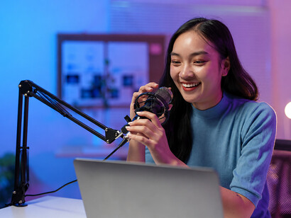 A podcast host speaks into a professional microphone in her home studio, using a laptop and creative workspace to produce digital content