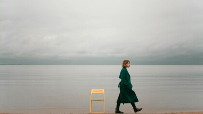 A woman in a green coat walks away from a chair by the ocean