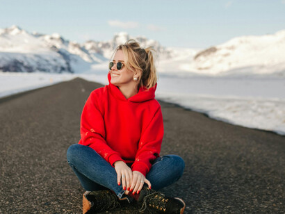 A stylish woman sits and poses on an empty road in Iceland, while a lone traveler walks away into the distance
