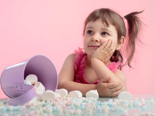 A cute little girl on a pink background, sitting at a table filled with marshmallows in front of her