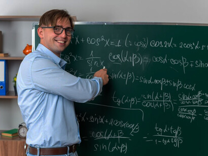 A young male mathematician wearing glasses stands beside a blackboard, writing equations and wiping them away