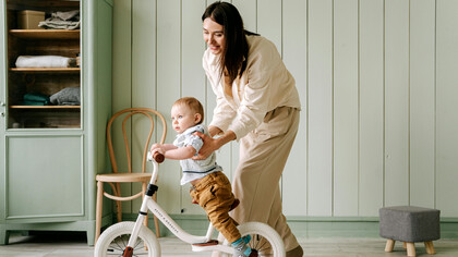 Mãe brinca com o seu filho bebê enquanto proporciona a ele as primerias experiências de estar em uma bicicleta infantil