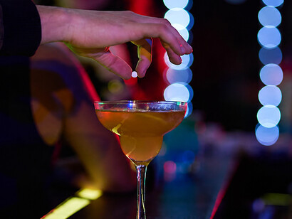 A man covertly placing drugs into a drink at a crowded bar, depicting drug-facilitated sexual assault