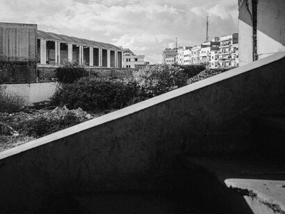 Vista urbana en blanco y negro tomada desde el interior de una estructura en ruinas por la guerra, encuadrando edificios residenciales e industriales al fondo