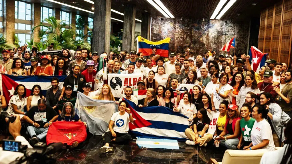 Integrantes da ALBA Movimentos reúnem-se durante a 4ª Assembleia Continental da organização, em Havana, diante das bandeiras de Cuba, Venezuela e Argentina. A imagem traduz a articulação política entre movimentos populares latino-americanos que o artigo apresenta como parte da disputa pela soberania e contra a ingerência imperial na região. A diversidade de movimentos representada na cena traduz o projeto de integração popular e solidariedade internacionalista que está no centro da articulação. A Alba Movimentos (Articulação Continental de Movimentos Sociais e Populares rumo à Alba) é uma rede que une centenas de organizações populares da América Latina e do Caribe — como sindicatos, grupos feministas, movimentos estudantis, camponeses e indígenas. Inspirada na aliança de governos da ALBA-TCP, ela atua de forma autônoma na base da sociedade para promover a integração regional e a resistência anticapitalista