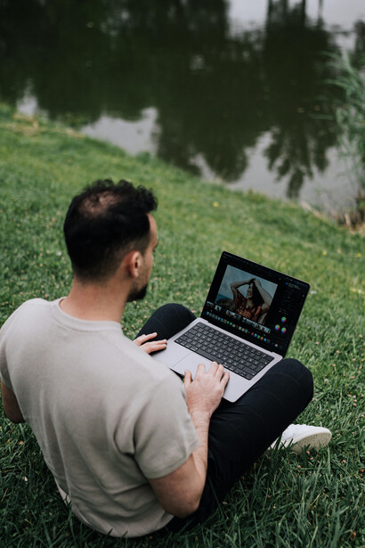 A man sits on the grass by a scenic pond in a park, using his laptop while working and enjoying the calm outdoor setting