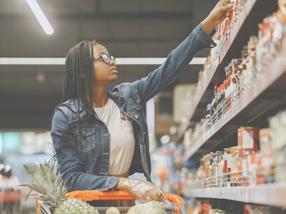 In a brightly lit supermarket, a young woman hesitates in front of a crowded shelf, visibly overwhelmed by the endless choices