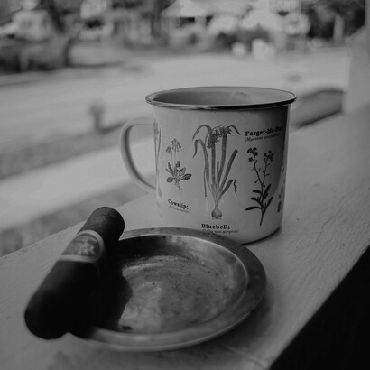 A rustic metal mug with botanical illustrations sits beside a cigar and an ashtray, embodying the contemplative rituals of cigar smoking